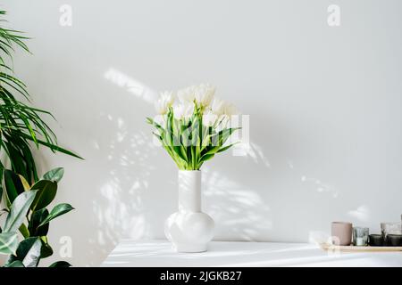 Skandinavische Innenausstattung mit Frühlingsstrauß aus weißen Tulpenblüten in Keramikvase, die auf dem Schrank steht. Minimalistisches Design mit grünen Pflanzen und Whi Stockfoto