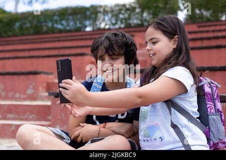 Lächelnder kaukasischer Junge und Mädchen von Grundschülerinnen, die vor dem Unterricht ein Foto mit einem Smartphone machen. Zurück zur Schule. Stockfoto