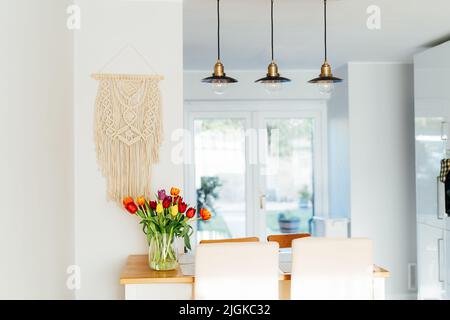 Stilvolle und moderne Boho, scandi Interieur der offenen Raum weiße Küche mit Tulpenblumen in Vase auf dem hölzernen Küchentisch, beige Macrame und Stockfoto