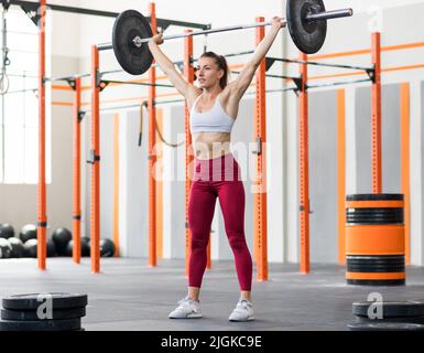 Eine körperstarke Sportlerin in Aktivbekleidung hält eine schwere Langhantel in den angehobenen Armen, während sie während des Gewichtheben-Trainings im Fitnessstudio eine Schnatterübung macht Stockfoto