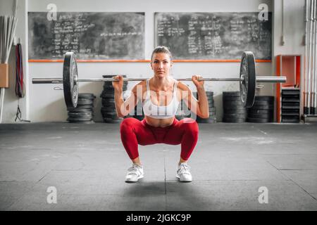 Fit weibliche Gewichtheber in Leggings und BH tun Langhantel zurück Hocke während funktionelles Training in geräumigen Fitnessraum tagsüber Stockfoto