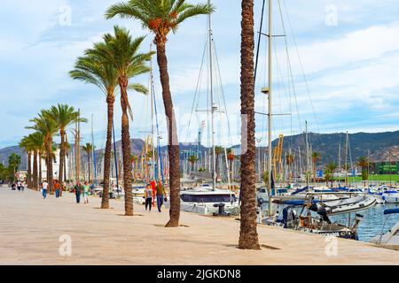 CARTAGENA, SPANIEN - 1. NOVEMBER 2021: Menschen, die am Ufer mit Palmen am Yachthafen mit Yachten spazieren, Cartagena, Spanien Stockfoto