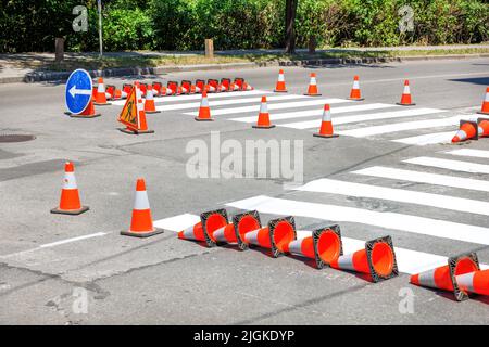 Anwendung von Markierungen auf einer Fußgängerüberfahrt über eine asphaltierte Straße. Frische Markierungen sind mit orangefarbenen Verkehrskegeln eingezäunt. Stockfoto
