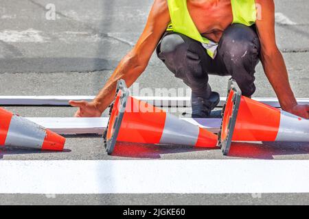 Ein Straßenarbeiter trägt mit einer Holzschablone und orangefarbenen Verkehrskegeln weiße Straßenmarkierungen auf einen Fußgängerübergang auf. Stockfoto