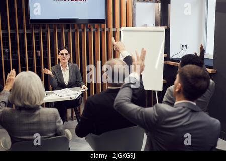 Selbstbewusste, attraktive Geschäftsfrau mit Brille, die am Tisch sitzt und Fragen von Geschäftseigentümern beantwortet, die an der Konferenz teilnehmen Stockfoto