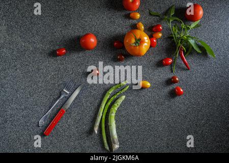 Lebensmittelrand mit grünem Spargel, roten und orangen Tomaten und rotem Chili mit Blättern. Gemüse, rotes Messer und Gabel auf dunkelgrauem Hintergrund t Stockfoto