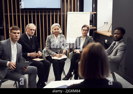 Gruppe von ernsthaften multiethnischen Geschäftsleuten unterschiedlichen Alters, die im Halbkreis gegen die Lehrerin sitzen und ihr aufmerksam beim Auffrischungskurs zuhören Stockfoto