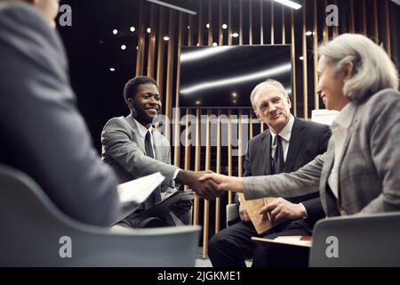 Positiv schöner junger afroamerikanischer Geschäftsmann, der im Kreis gleichgesinnter Menschen sitzt und mit einer asiatischen Dame handshake, während er neue Leute trifft Stockfoto