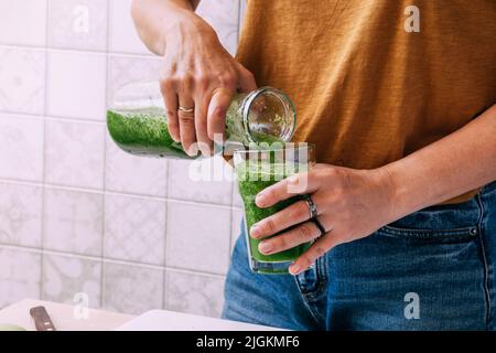 Eine Frau gießt einen frisch zubereiteten Smoothie aus grünem Gemüse und Obst in Glas. Vegetarismus Stockfoto