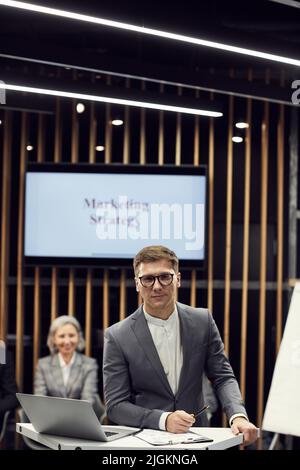 Content-Portrait hübscher junger Vermarkter in Brillen, der am Sprechtisch mit Zwischenablage und Laptop beim Business Forum steht Stockfoto