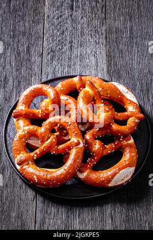 Weiche Brezeln in Form von Knoten gebacken und mit Salz auf schwarzem Teller auf dunklem Holztisch bestreut, vertikale Ansicht von oben, freier Platz Stockfoto