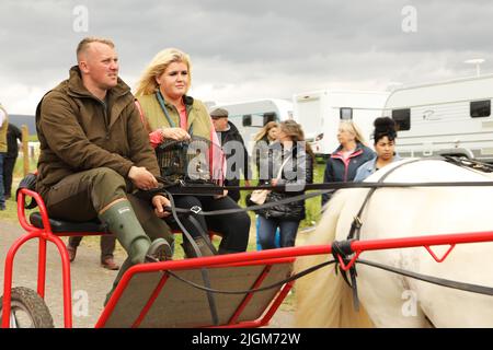 Ein graues Pferd, das ein Paar in eine Falle zieht, eine Frau, die einen Vogelkäfig trägt. Appleby Horse Fair, Appleby in Westmorland, Cumbria, England, Vereinigtes Königreich Stockfoto