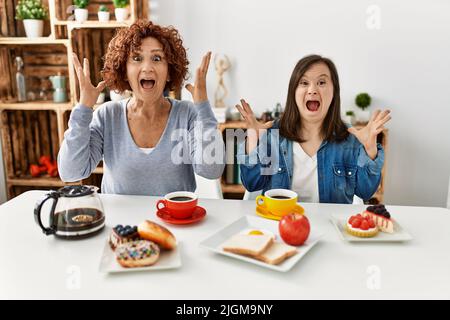 Familie von Mutter und Down-Syndrom Tochter zu Hause sitzen Frühstück feiern verrückt und erstaunt für den Erfolg mit erhobenen Armen und offenen Augen Stockfoto