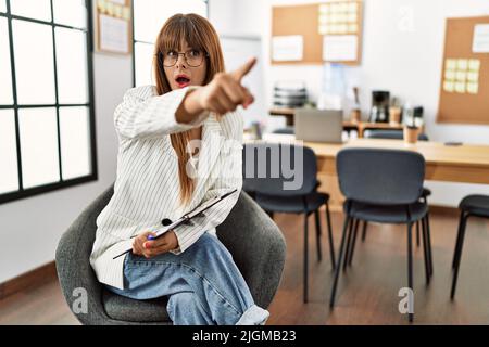 Hispanische Geschäftsfrau, die im Büro arbeitet, zeigt mit dem Finger überrascht voraus, offener Mund verblüfft, etwas auf der Vorderseite Stockfoto