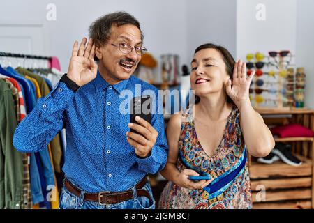 Interrassisches Paar im mittleren Alter im Einzelhandelsgeschäft mit einem Smartphone, das lächelt und mit der Hand über das Ohr einem Gerücht oder Klatsch lauscht. Taubheitskonzept. Stockfoto