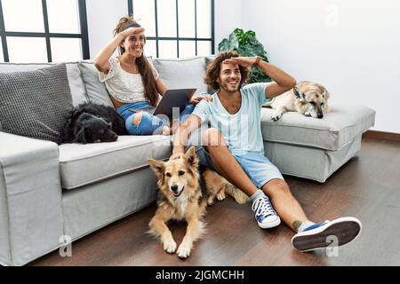 Junge hispanische Paar mit Hunden entspannen zu Hause sehr glücklich und lächelnd Blick weit weg mit Hand über Kopf. Suchkonzept. Stockfoto