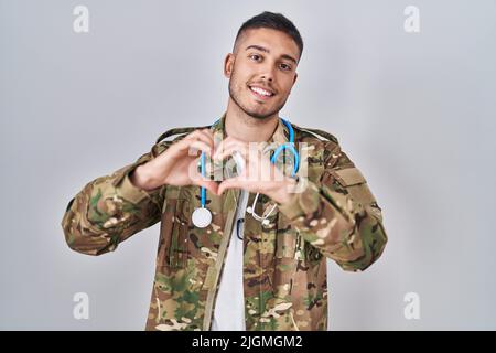 Junge hispanische Arzt trägt Tarnung Armee Uniform lächelnd in der Liebe tun Herz Symbol Form mit Händen. Romantisches Konzept. Stockfoto