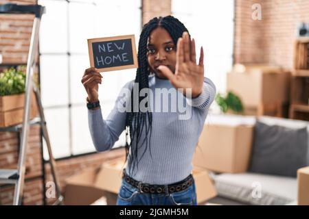 afroamerikanische Frau hält Tafel mit neuem Heimattext mit offener Hand und tut Stoppschild mit ernsthaftem und selbstbewusster Ausdruck, Verteidigungsgeste Stockfoto