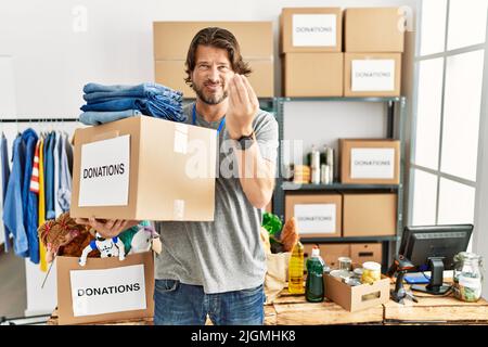 Ein gutaussehender Mann mittleren Alters, der am Freiwilligenstand eine Spendenbox für wohltätige Zwecke hält und mit Hand und Fingern eine italienische Geste mit selbstbewusster Ausdruckskraft macht Stockfoto