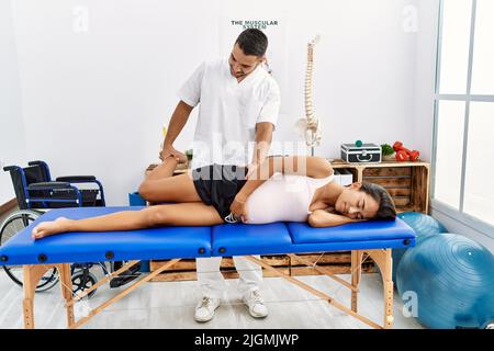 Lateinischer Mann und Frau in Physiotherapeut-Uniform mit Schwangerschaftsrehabsession Stretching Bein in Physiotherapie-Klinik Stockfoto