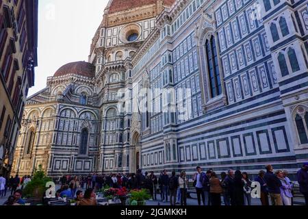 Leute in der Schlange, entlang der Seite der großen Kathedrale, warten darauf, den Dom zu betreten, während andere an Restauranttischen im Freien sitzen, Florenz, Italien. Stockfoto