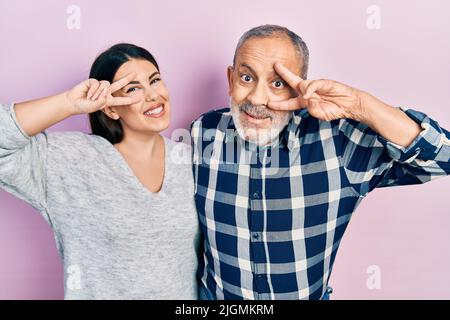 Hispanische Vater und Tochter tragen legere Kleidung und tun das Friedenssymbol mit den Fingern über dem Gesicht, lächeln fröhlich und zeigen den Sieg Stockfoto