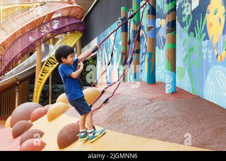 Glücklicher asiatischer Junge, der Seil zieht, um auf dem Spielplatz im Stadtpark zu klettern. Kind spielt nach dem Unterricht im Kindergarten im Freien. Stockfoto