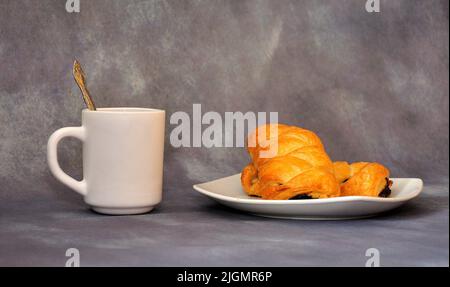 Ein Teller mit zwei Blätterteiggebäck mit Mohnsamen und einer Tasse heißen Kaffee auf einem grauen abstrakten Hintergrund. Nahaufnahme. Stockfoto