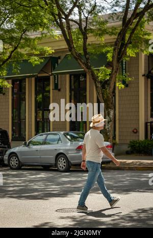 Ein Mann in einem urbanen, zwanglosen Lebensstil, der einen Bürgersteig entlang der Straße in einer Stadt entlang läuft. Stilvoller Mann in einem T-Shirt, einer Jeans aus blauem Denim und einem Hut, der in einem Stockfoto