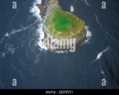 Blick von oben auf eine kleine Insel im Meer mit grünem Gras bedeckt. Die weißen Wellen des blauen Ozeans waschen die Insel von allen Seiten. Schöne Natur, Stockfoto