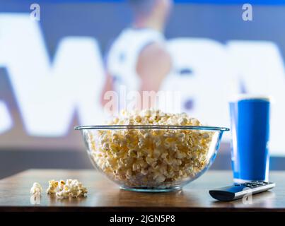 Vor dem Hintergrund eines Basketballspiels auf dem Bildschirm eines großen Plasma-Fernsehers, Popcorn in einer Glasschale, einer Fernbedienung und einem Getränk in einem gla aus Kunststoff Stockfoto