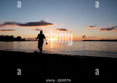 Mann, der bei Sonnenuntergang am Strand Fußball spielt. Dunkler Blick am Abend. Fußballspieler Silhouette auf einem Sandstrand am späten Abend. Stockfoto