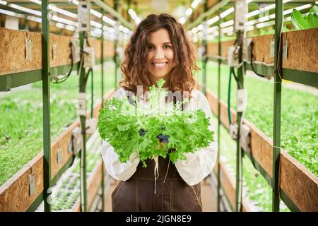 Fröhliche Gärtnerin, die die Kamera anschaut und lächelt, während sie die grüne Blattpflanze hält. Junge Frau, die im Gang zwischen Regalen mit Pflanzen im Gewächshaus steht. Stockfoto