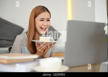 Hübsche und fröhliche junge asiatische Lebensmittelbloggerin Influencerin im Café, die ihren Online-Followern per Laptop einen leckeren Kuchen vorführt. Stockfoto