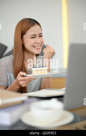 Glückliche, tausendjährige asiatische Lebensmittelbloggerin Influencer im Café, die ihren Online-Followern über einen Laptop einen leckeren Kuchen vorführt. Stockfoto