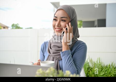 Schöne und elegante, tausendjährige asiatische muslimische Frau, die Hijab trägt und mit jemandem im Hinterhof des Hauses telefoniert. Stockfoto