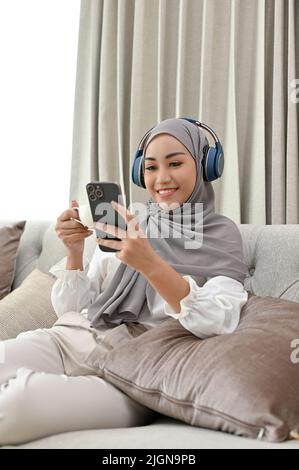 Porträt einer eleganten, tausendjährigen asiatischen Muslimin in Hijab und Kopfhörern, die Musik hört, während sie in ihrer komfortablen Livi einen heißen Kaffee schlürft Stockfoto