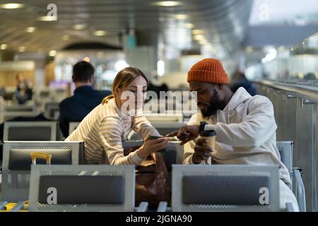 Junges internationales Paar, das im Flughafenterminal auf das Einsteigen wartet, mit dem Mobiltelefon Stockfoto
