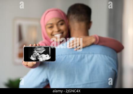 Happy Islamic Lady Zeigt Smartphone Mit Baby Ultraschall-Bild Und Umarmen Mann Stockfoto