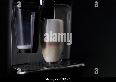 Frisch gebrühter Latte wird aus der Kaffeemaschine in Glasbecher gegossen. Professionelles Brühen von Kaffee zu Hause. Stockfoto