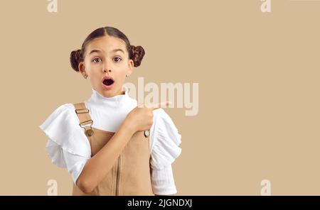 Verblüfft verblüfft betäubt präteen Mädchen mit Finger zeigt auf Werbeprodukt auf beige Hintergrund. Stockfoto