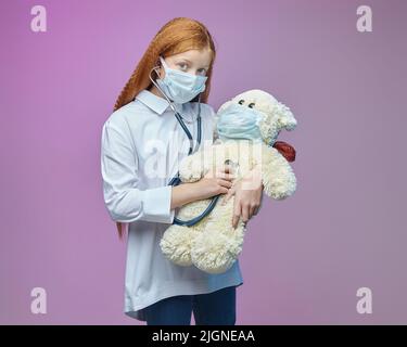 Kleines Mädchen in einer medizinischen Maske hört einem Teddybär durch ein Stethoskop zu. Das Konzept der Prävention covid-19 Stockfoto