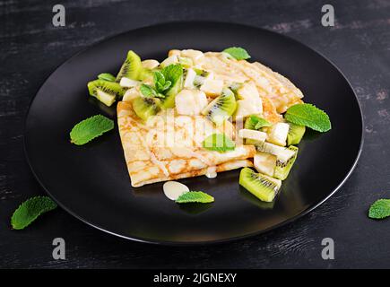 Crepes, dünne Pfannkuchen oder Blini mit Kiwi und Banane auf schwarzem Teller. Pfannkuchen Woche. Shrovetide. Stockfoto