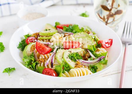 Fusilli-Nudelsalat mit Avocado, Tomaten, frischem grünem Salat, roten Zwiebeln und Senfdressing auf weißem Hintergrund. Vegetarisch gesundes Mittagessen. Stockfoto