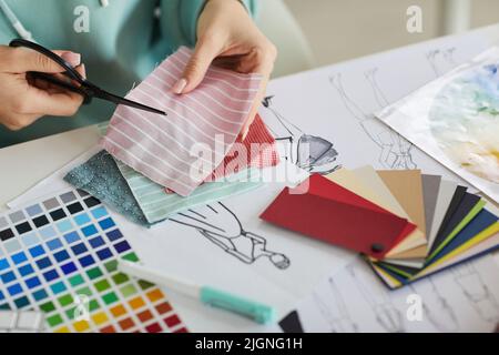 Nahaufnahme einer nicht erkennbaren Frau, die mit Paletten und Modezeichnungen am Schreibtisch sitzt und im Schneideratelier Stoff schneidet Stockfoto