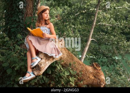 Niedliches kaukasisches Schulmädchen in einem Kleid und einem Strohhut, das mit einem Buch auf einem alten Holzbuch posiert. Im Hintergrund sind Park und Fluss. Das Konzept von sch Stockfoto