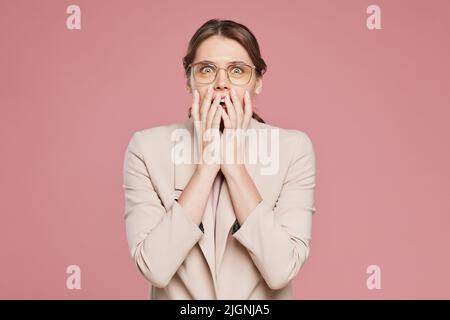 Porträt einer verblüfften attraktiven jungen Frau in Jacke und Brille, die vor rosa Hintergrund steht und den Mund mit Händen in Aufregung bedeckt Stockfoto