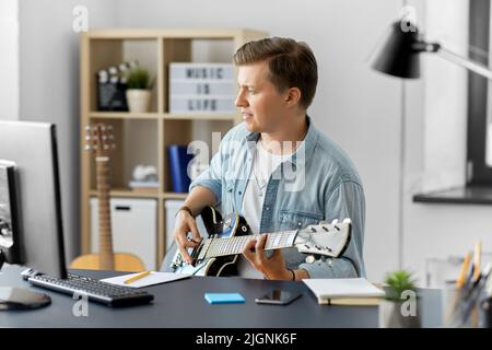 Junger Mann mit Computer, der zu Hause Gitarre spielt Stockfoto