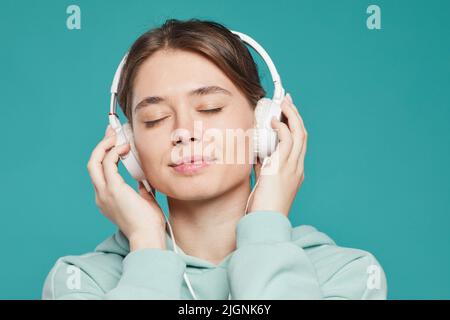 Ruhige, attraktive junge Frau mit geschlossenen Augen, die Kopfhörer mit Kabelanschluss trägt und entspannende Musik vor blauem Hintergrund hört Stockfoto