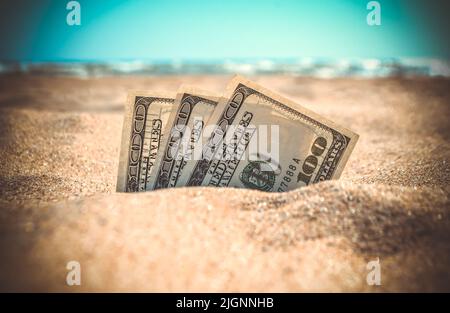 Die mit Sand bedeckten Gelddolare liegen am Sandstrand in der Nähe der Meereswellen am sonnigen Sommertag aus nächster Nähe. Geld wächst aus dem Boden. Konzept Finanzierung Geld Urlaub Relax Urlaub. Stockfoto